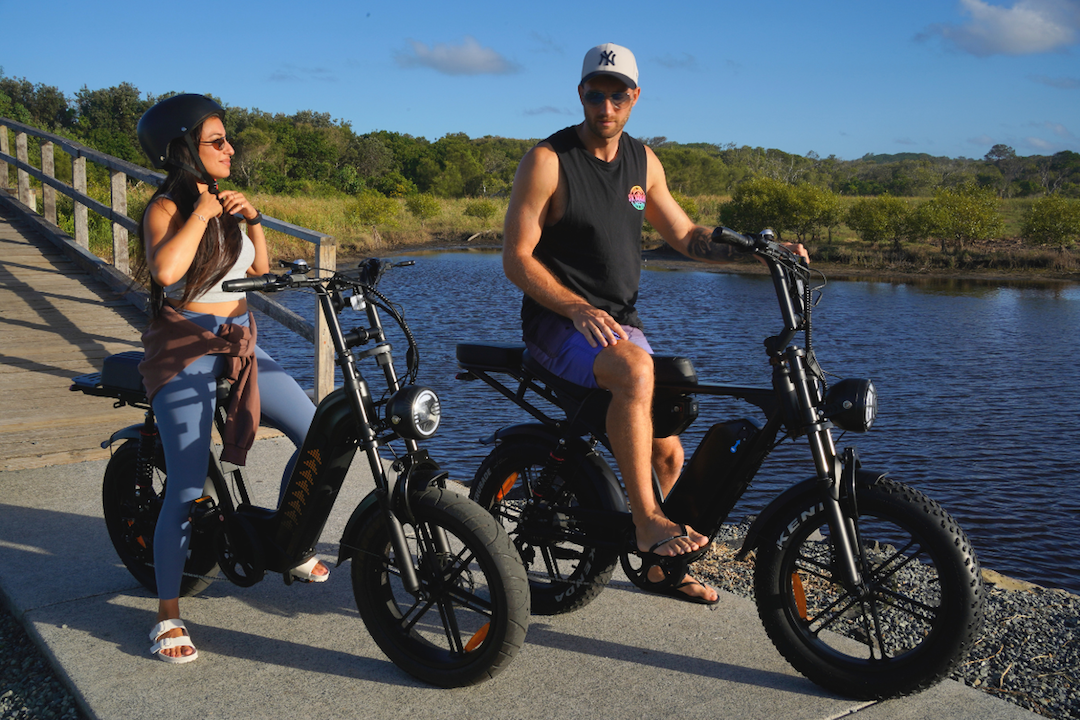 Couple riding fat tire eBikes on a scenic waterfront path in sunny weather.
